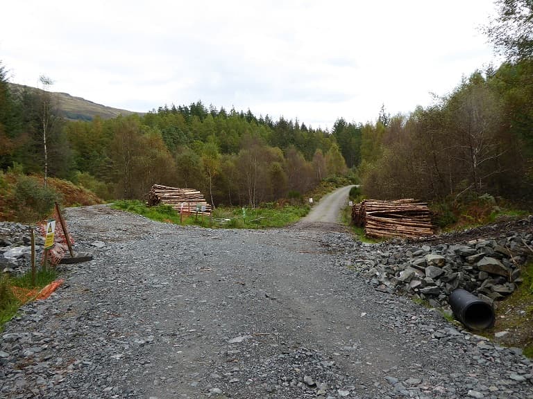 A forestry track of loose stone meeting a surfaced road, with stacked cut timber on both sides and a barrier across the junction.